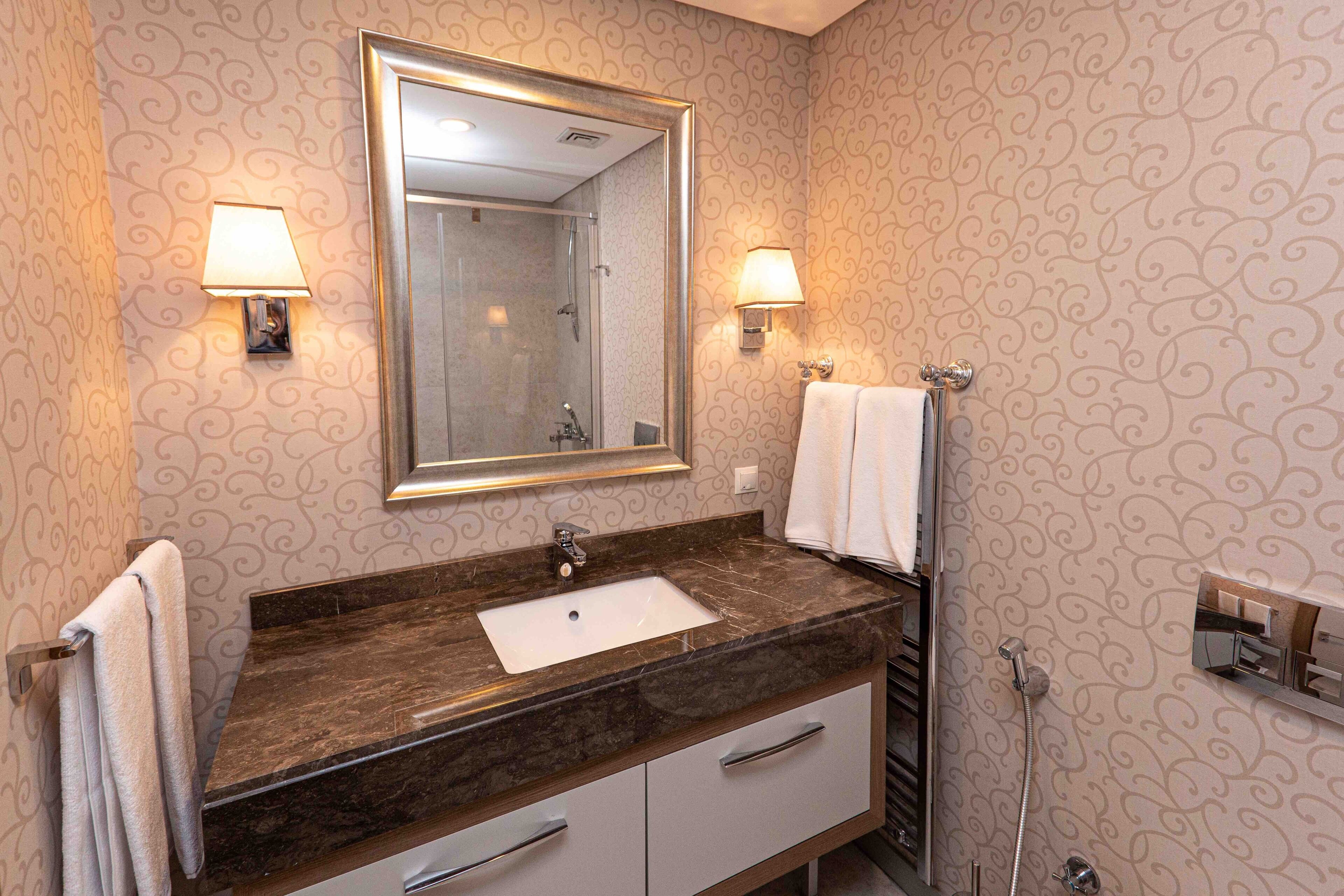 Un baño con lavamanos de mármol, dos lámparas de pared, un espejo grande y un perchero para toallas.