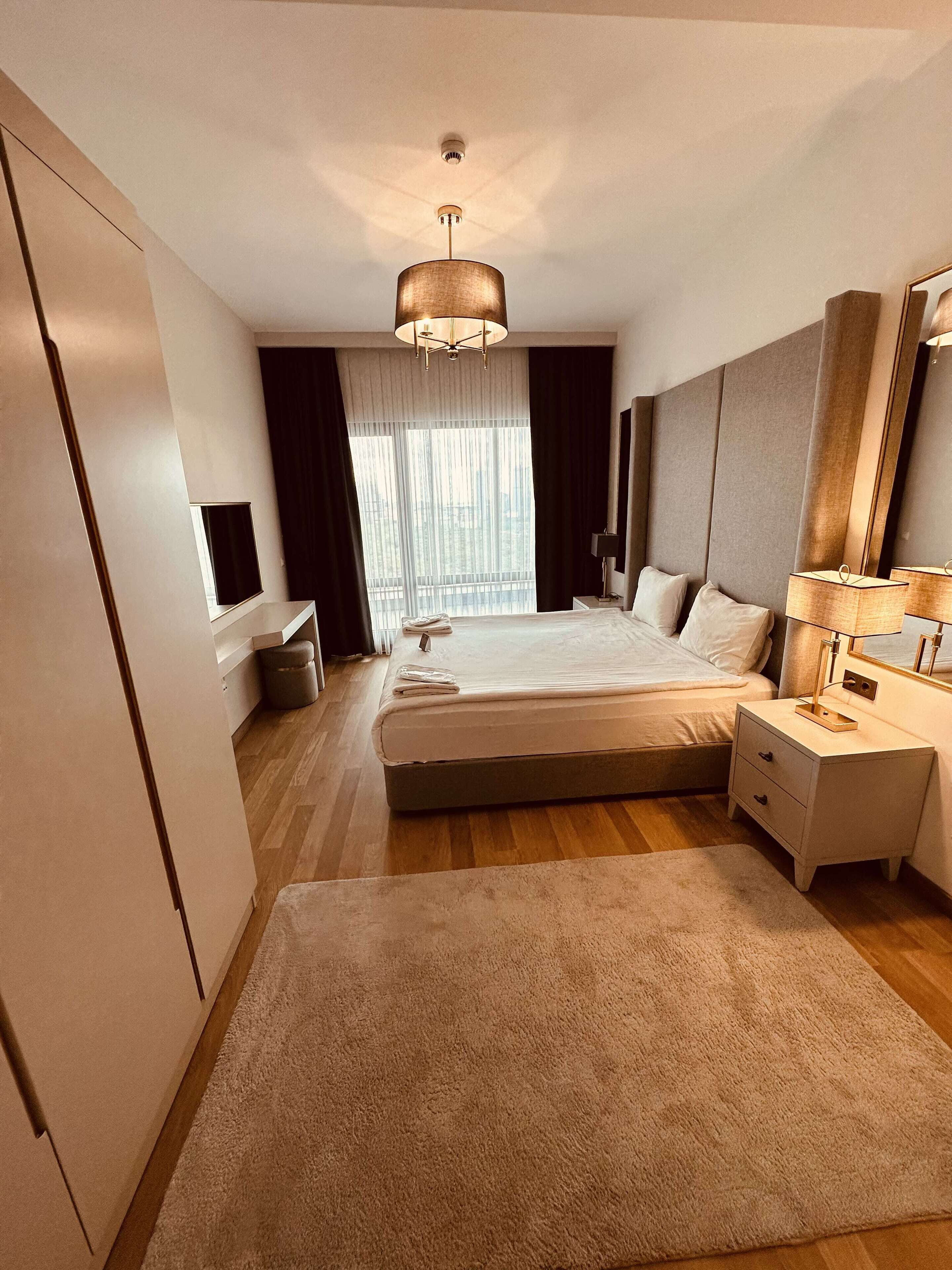 A modern hotel room with a large bed, bedside tables, a desk, and a large window with curtains.