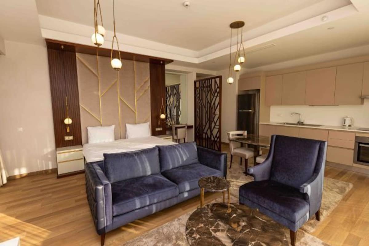 Una habitación de hotel moderna con una cama, un sofá azul, una mesita de centro redonda y una zona de comedor con una cocina al fondo.