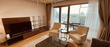 A modern living room with a flat-screen TV, a marble coffee table, two armchairs, a sofa, and a large window with a city view.