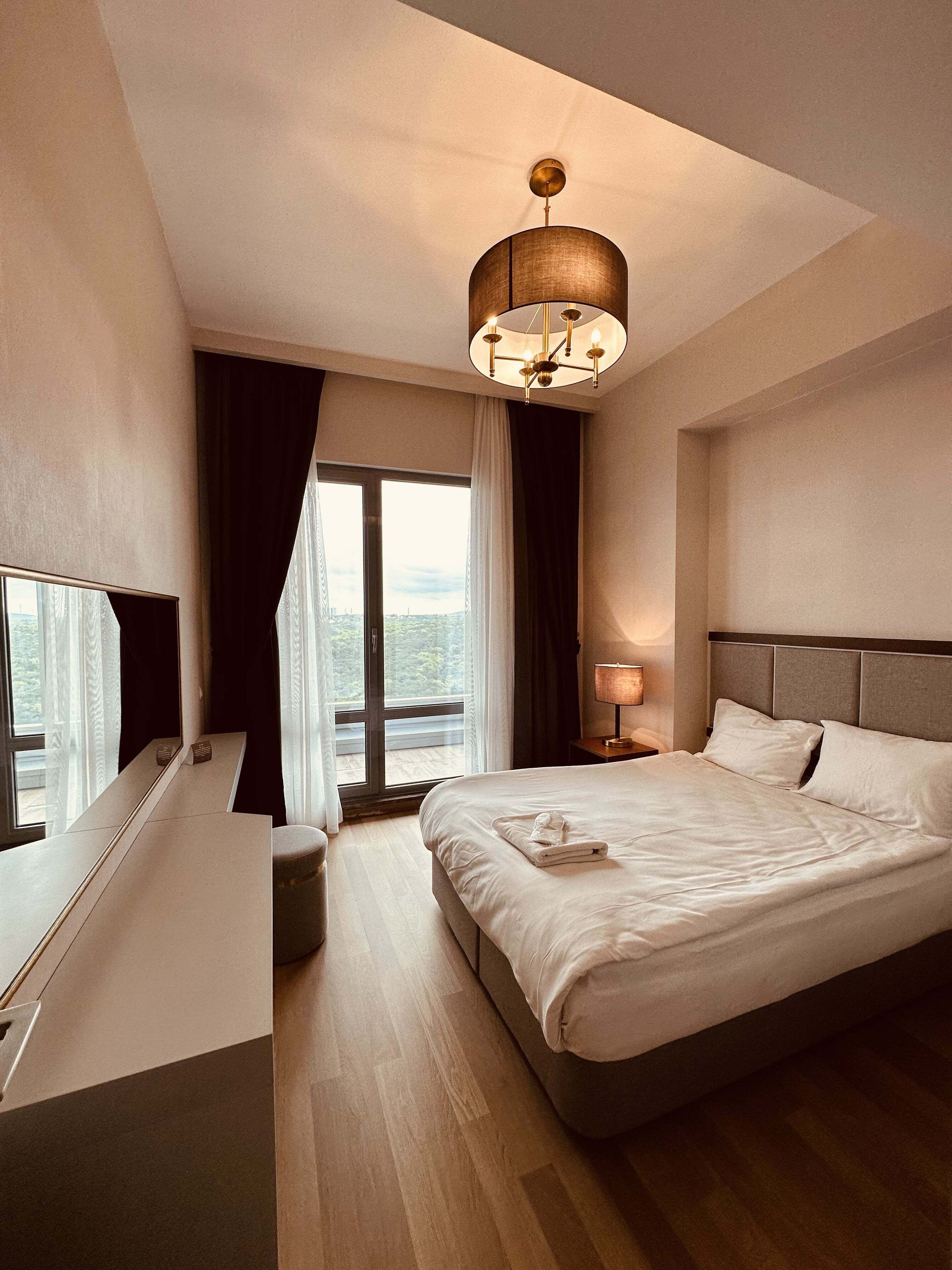 A modern hotel room with a large bed, a desk, a chair, a nightstand, a lamp, and a view of the city through the window.