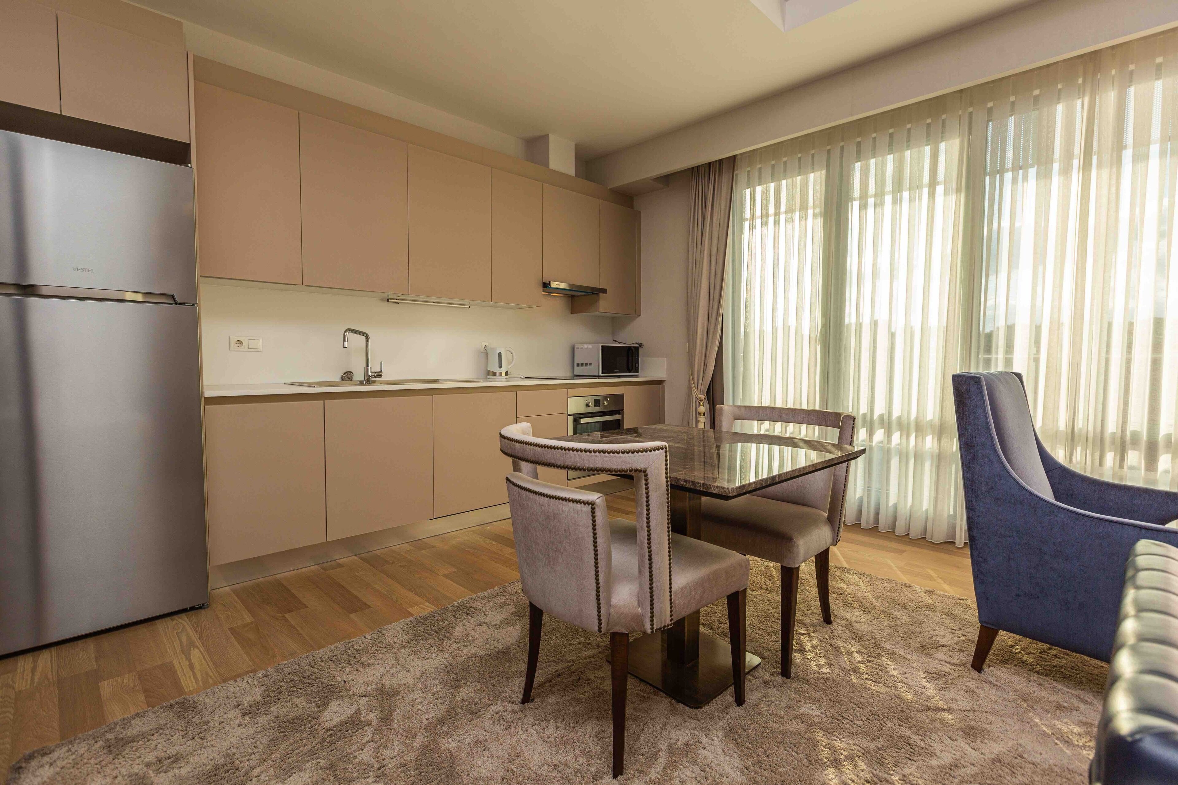 A modern kitchen with stainless steel appliances, a dining table with chairs, and a large window with sheer curtains.