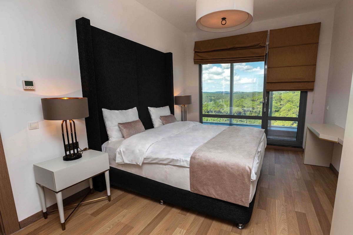 Une chambre moderne avec un grand lit, deux tables de chevet, un bureau et une vue sur un paysage verdoyant par la fenêtre.