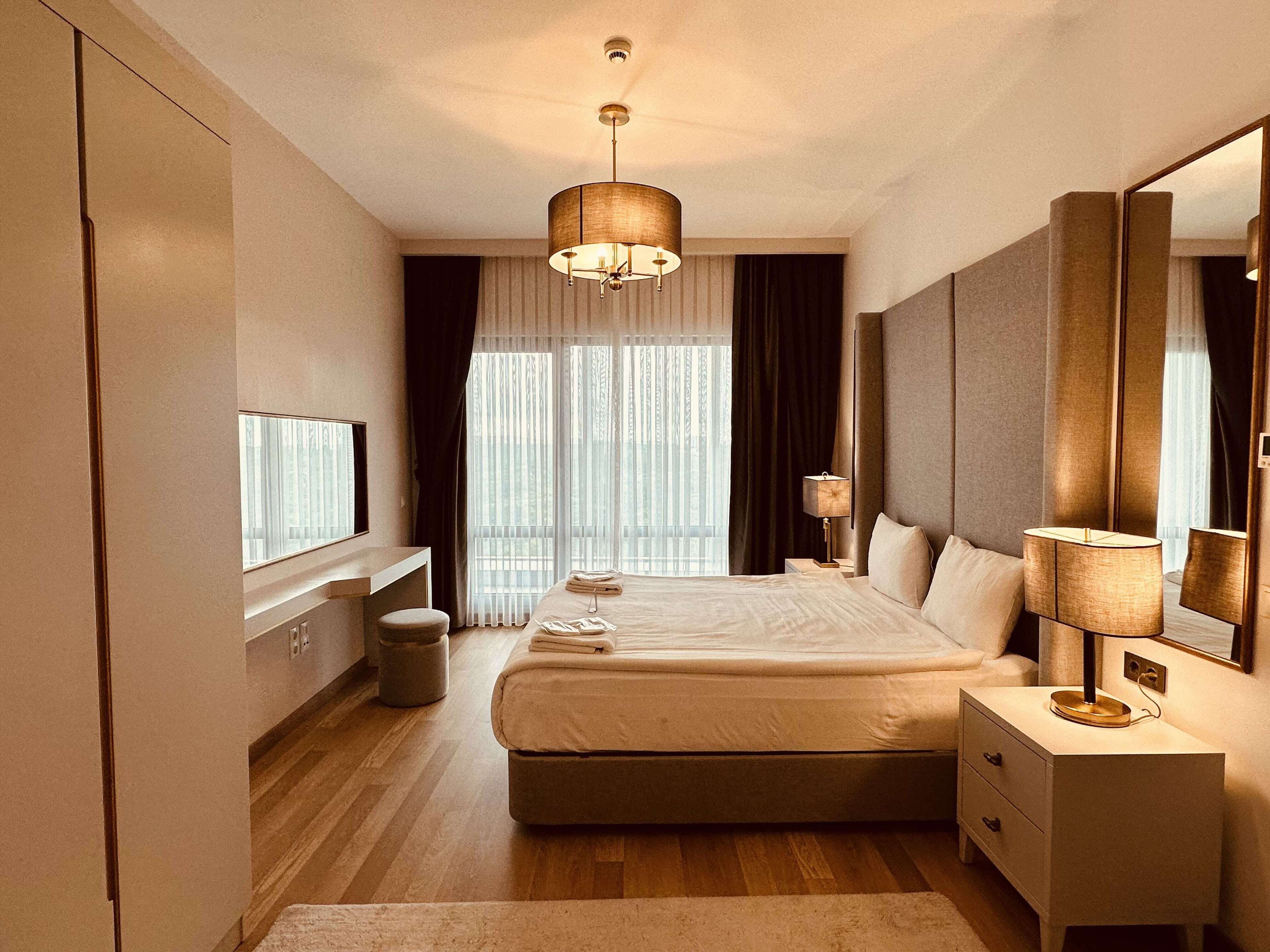 A modern hotel room with a large bed, bedside tables, a mirror, and a window with curtains.
