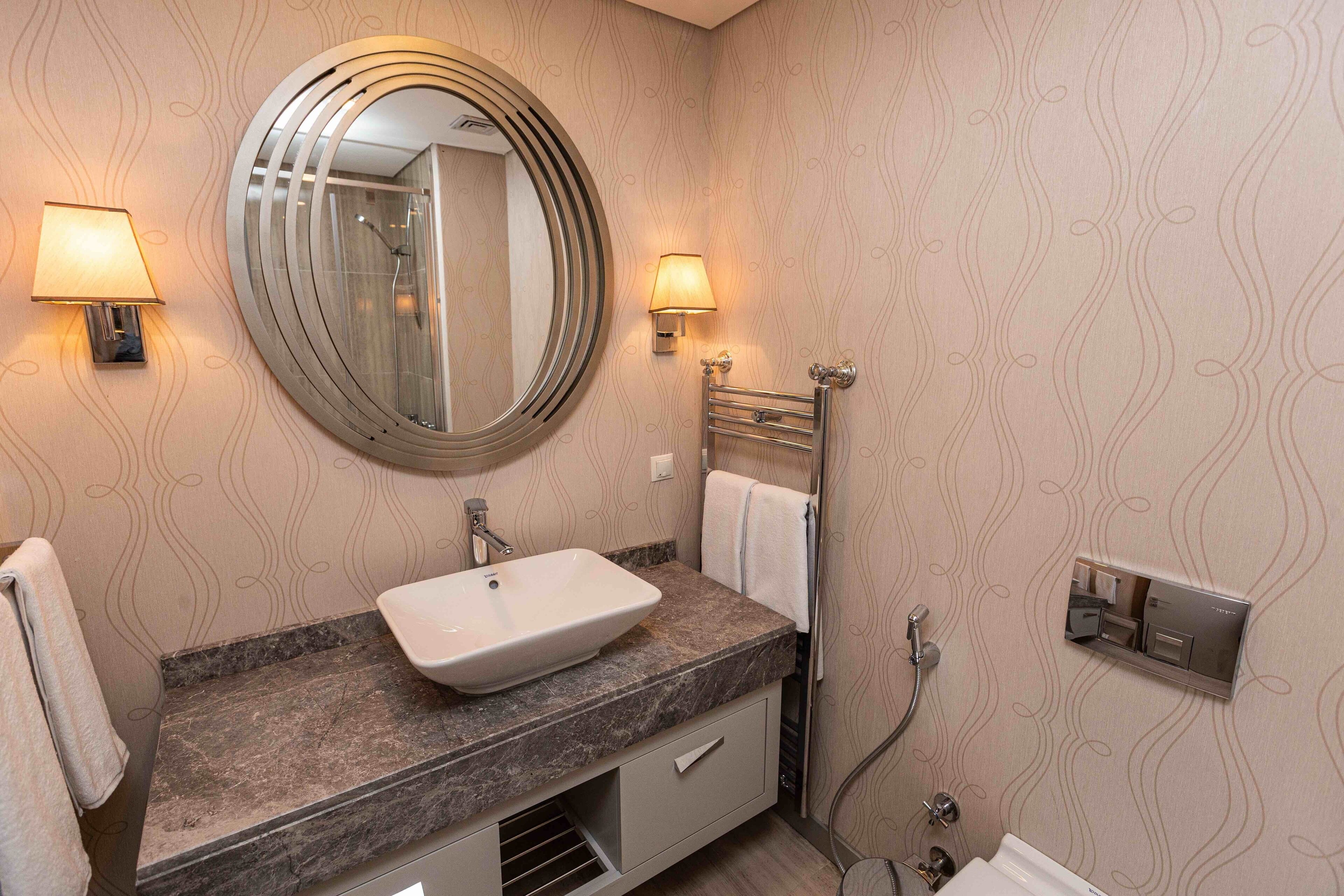 A bathroom with a round mirror, two wall-mounted lamps, a sink, and a towel rack.