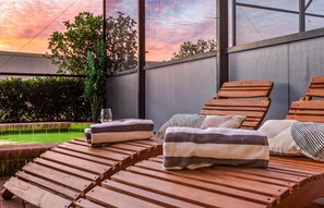 Ein sonniger Außenbereich mit Holzliegen, einem Glastisch und Blick auf einen Pool und Bäume.