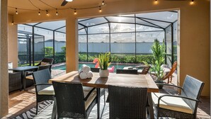 Um alpendre coberto com mesa e cadeiras, ventoinha no teto, luzinhas e vista para uma piscina e montanhas.