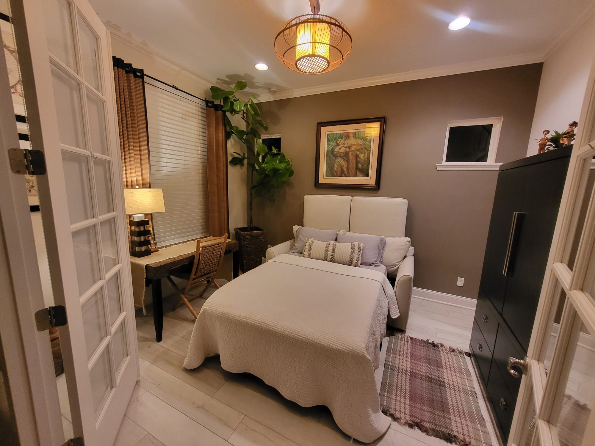A bedroom with a bed, bedside table, lamp, curtains, a plant, a painting, and a wardrobe.