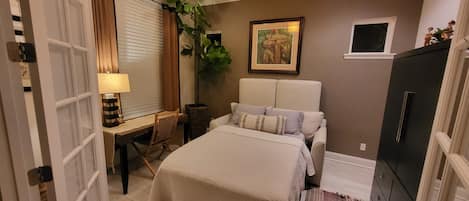 A bedroom with a bed, bedside table, lamp, curtains, a plant, a painting, and a wardrobe.