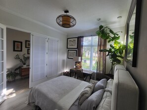 A bedroom with a bed, a desk, a chair, a window with curtains, and a plant.