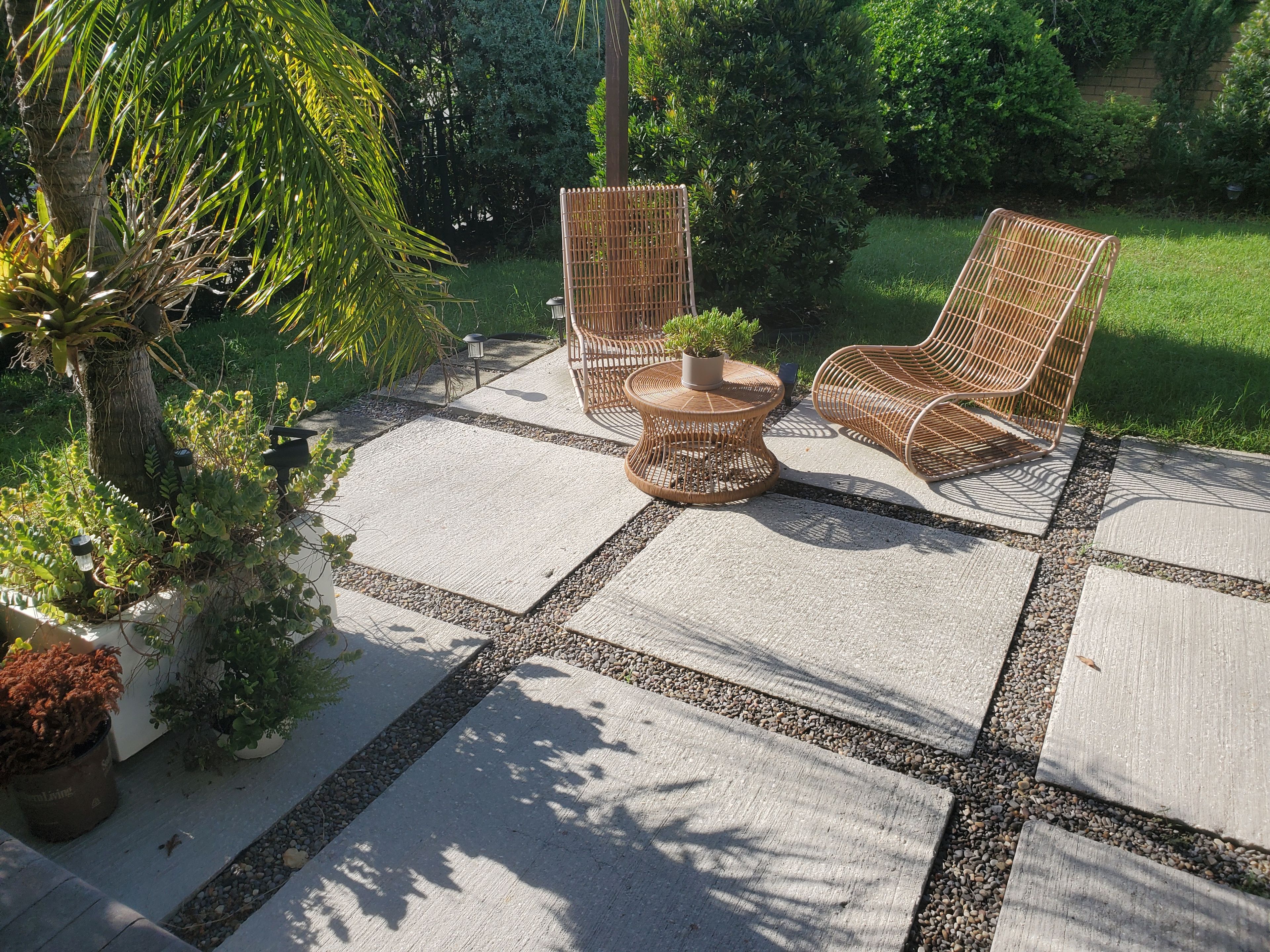 A patio area with a concrete floor, two wicker chairs, a small round table with a potted plant, and surrounding greenery.
