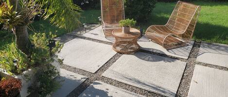 A patio area with a concrete floor, two wicker chairs, a small round table with a potted plant, and surrounding greenery.