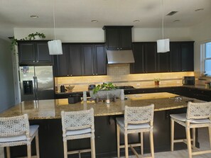 A modern kitchen with dark cabinetry, a stainless steel refrigerator, a large island with bar stools, and pendant lighting.