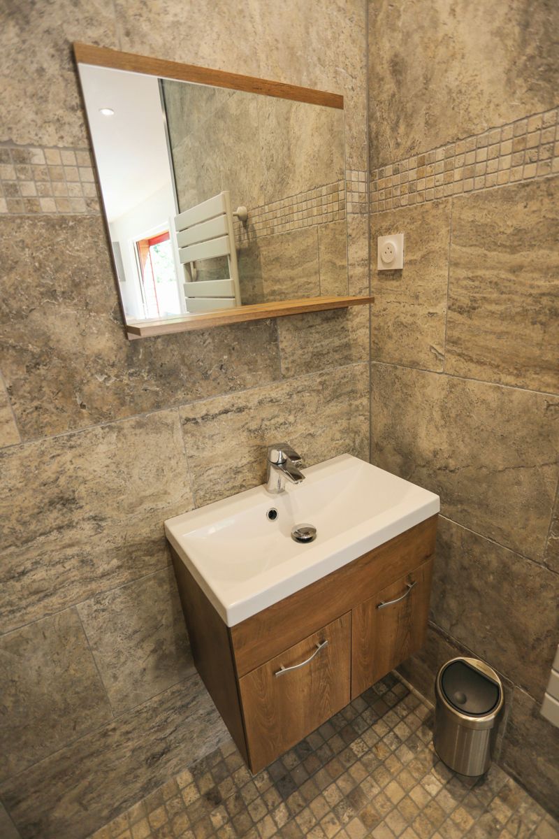 A bathroom with a stone wall, a white sink, wooden cabinets, a mirror, and a trash can.