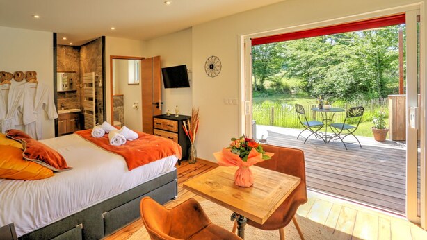 Een slaapkamer met een bed, een houten tafel en een stoel, een badkamer met een spiegel, en uitzicht op een tuin door een geopende deur.