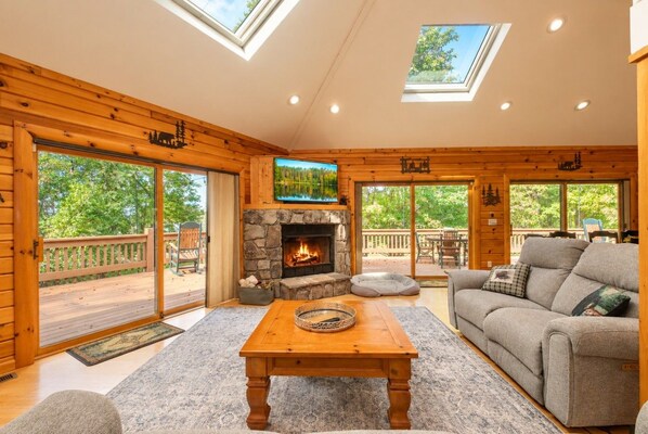 A log cabin interior with a stone fireplace, wooden furniture, and large windows.