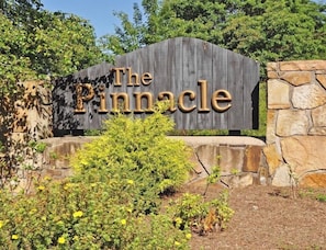 A wooden sign with the text 'The Pinnacle' in a rustic font, surrounded by greenery and a stone wall.