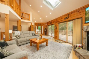 A spacious living room with a wooden ceiling, a fireplace, and a large window overlooking a deck.