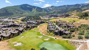 Un campo de golf con un club de campo, rodeado de edificios residenciales y montañas al fondo.