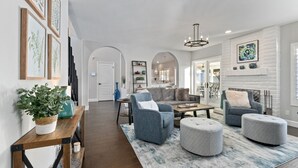 Un salon moderne avec un canapé, des poufs, une table basse et une cheminée. Des tableaux encadrés ornent le mur, et une plante est posée sur une table d’appoint.