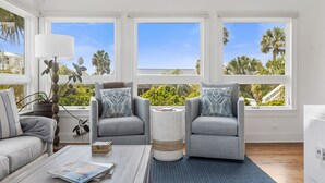 Una sala moderna con dos sillones grises, una mesa de centro y una lámpara. Amplias ventanas ofrecen vistas a palmeras y un edificio.