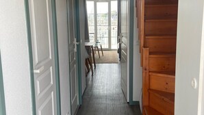 A hallway with a wooden staircase, a view of the outside through a glass door, and a small table with chairs. - Port du Crouesty. duplex rénové, calme, terrasse, idéal en famille (6 pers) (Arzon)