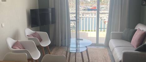 Een moderne woonkamer met een bank, een glazen salontafel en een eettafel met stoelen. Er is een balkon met uitzicht op boten en gebouwen.