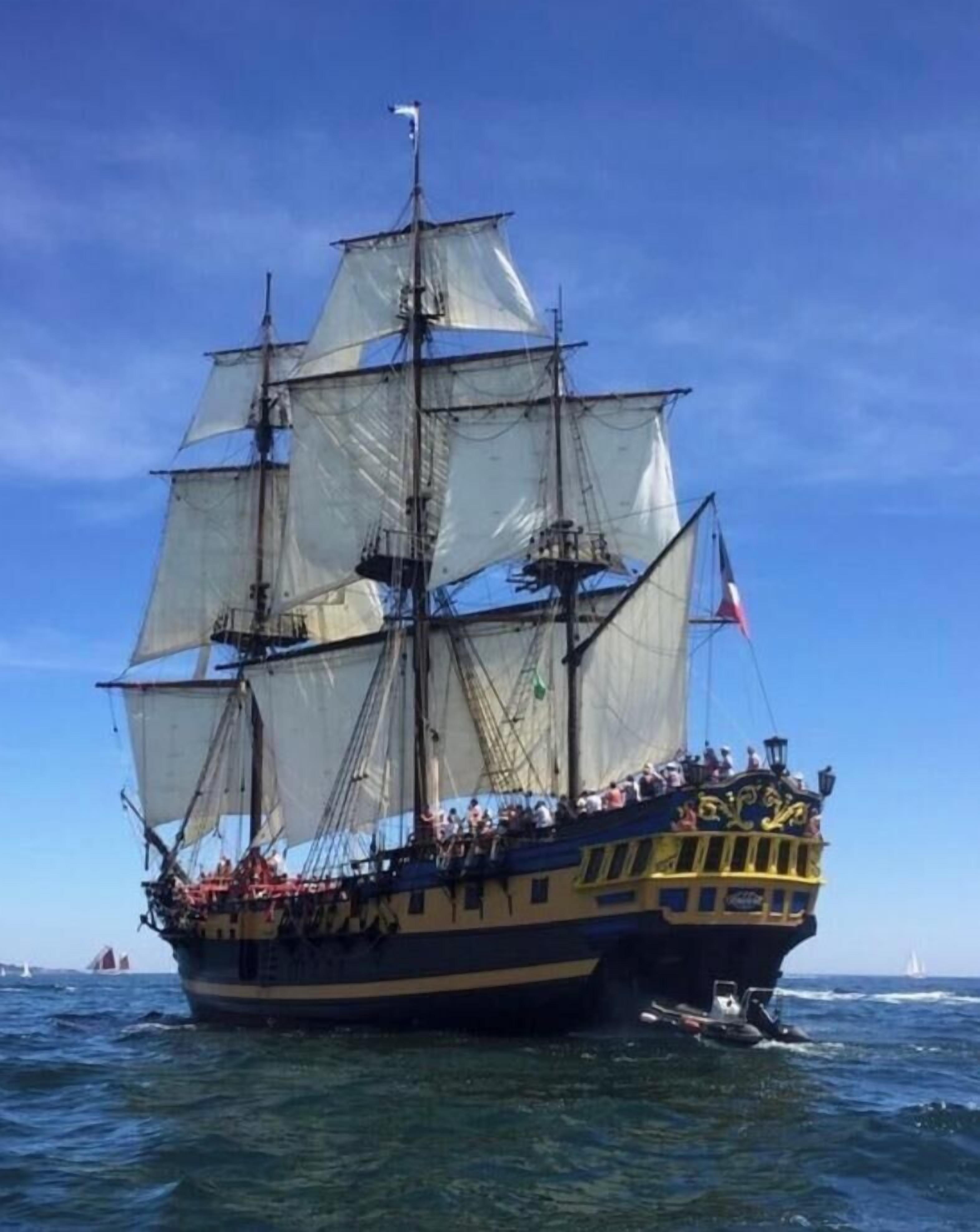 Een groot zeilschip met meerdere masten en zeilen, vol passagiers, op het water.