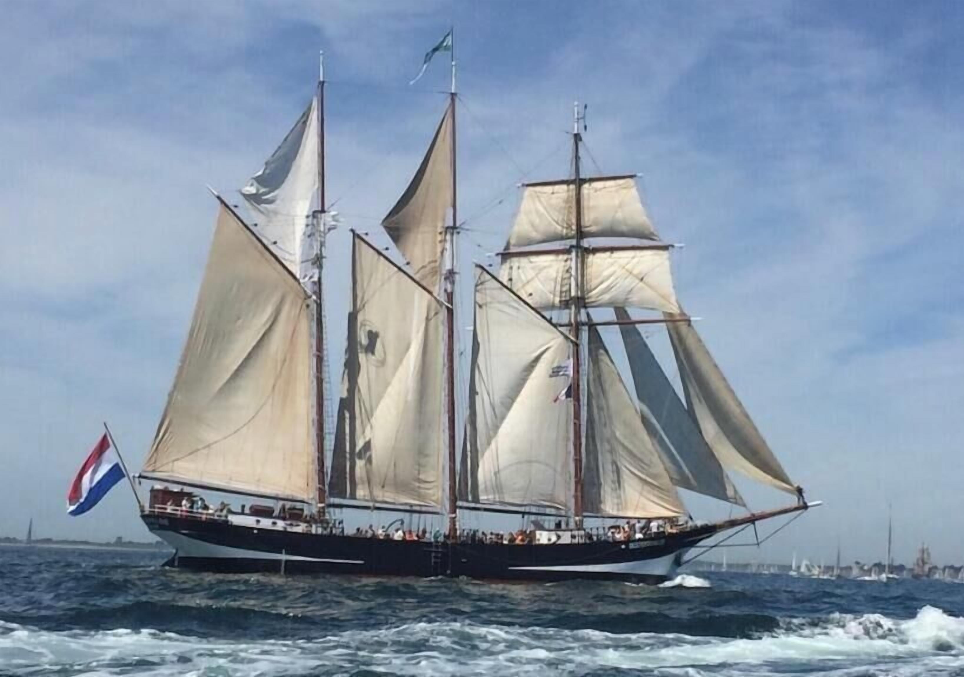 Ein Segelschiff mit mehreren Masten und Segeln, Passagiere auf dem Deck und einer Flagge.