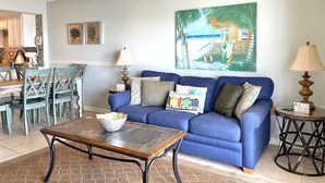 A living room with a blue sofa, a coffee table, and a dining area with chairs and a table. - Surf Side Shores 1906 | 3 bd 3 ba | gulf-front  (Gulf Shores)
