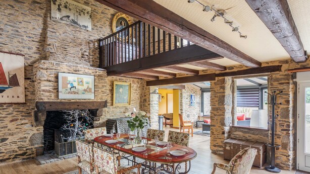A spacious room with a stone fireplace, a wooden staircase, and a dining area with floral chairs and a long table. - LA LONGERE DU TERTRE - Charmante longère proche plage (Cancale)