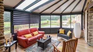 A spacious room with a red leather sofa, a black wicker sofa, and a wooden chair. There is a table with a lamp and a tray of pastries. The room has large windows with blinds and a view of the garden outside. - LA LONGERE DU TERTRE - Charmante longère proche plage (Cancale)