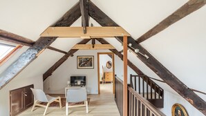 An attic room with exposed wooden beams, a desk with a chair, a television, and a staircase leading to another level. - LA LONGERE DU TERTRE - Charmante longère proche plage (Cancale)