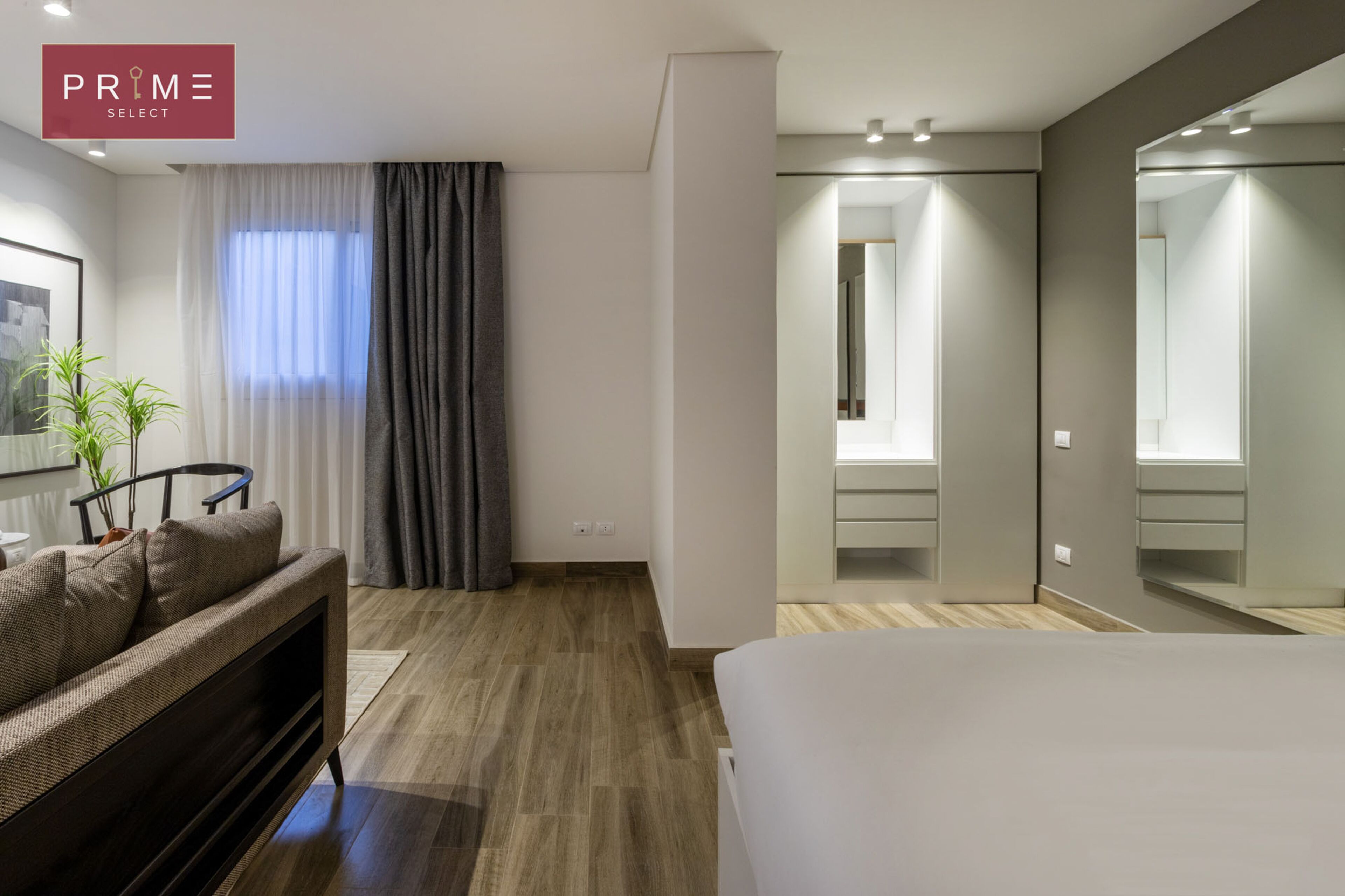 Ein modernes Hotelzimmer mit Holzboden, einem Bett, einer Couch, einem Spiegel und einem fenster mit Vorhängen.
