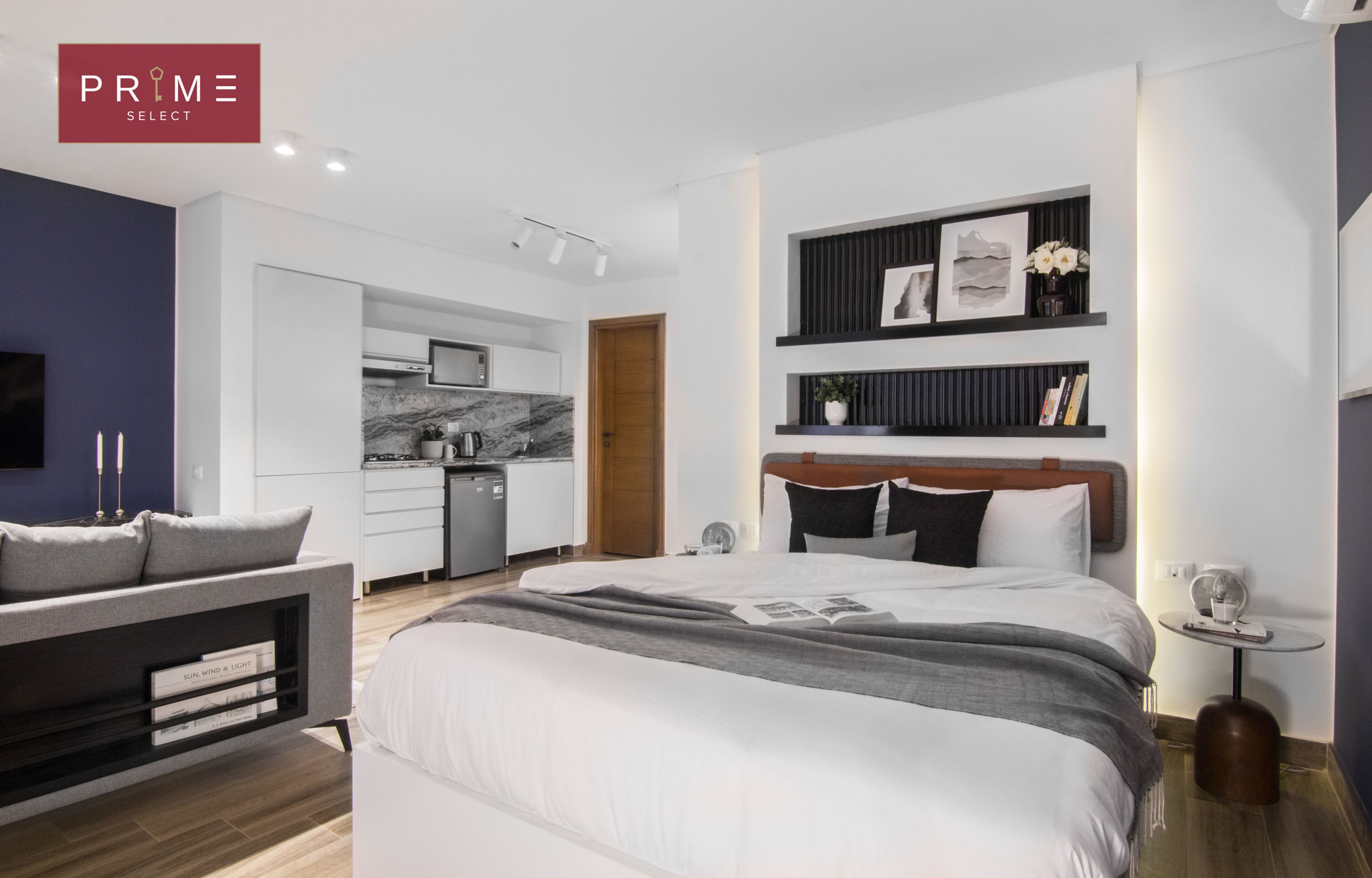 Ein modernes Schlafzimmer mit einem großen Bett, Nachttischen, einem fest installierten Fernseher und einer kleinen Küchenzeile.