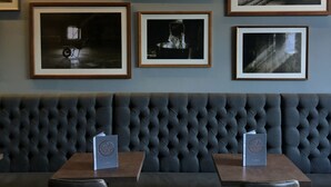 A restaurant interior with a dark blue upholstered bench, framed black and white photographs on the wall, and two tables with speakers. - Kirkwall Hotel (Kirkwall)