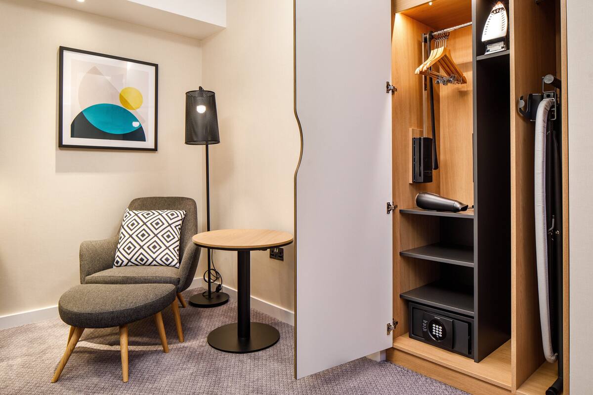Ein modernes Zimmer mit grauem Sessel, einem kleinen runden Tisch, einem gerahmten abstrakten Kunstwerk, einer Stehlampe und einem begehbaren Kleiderschrank mit Regalen und Kleiderstangen.