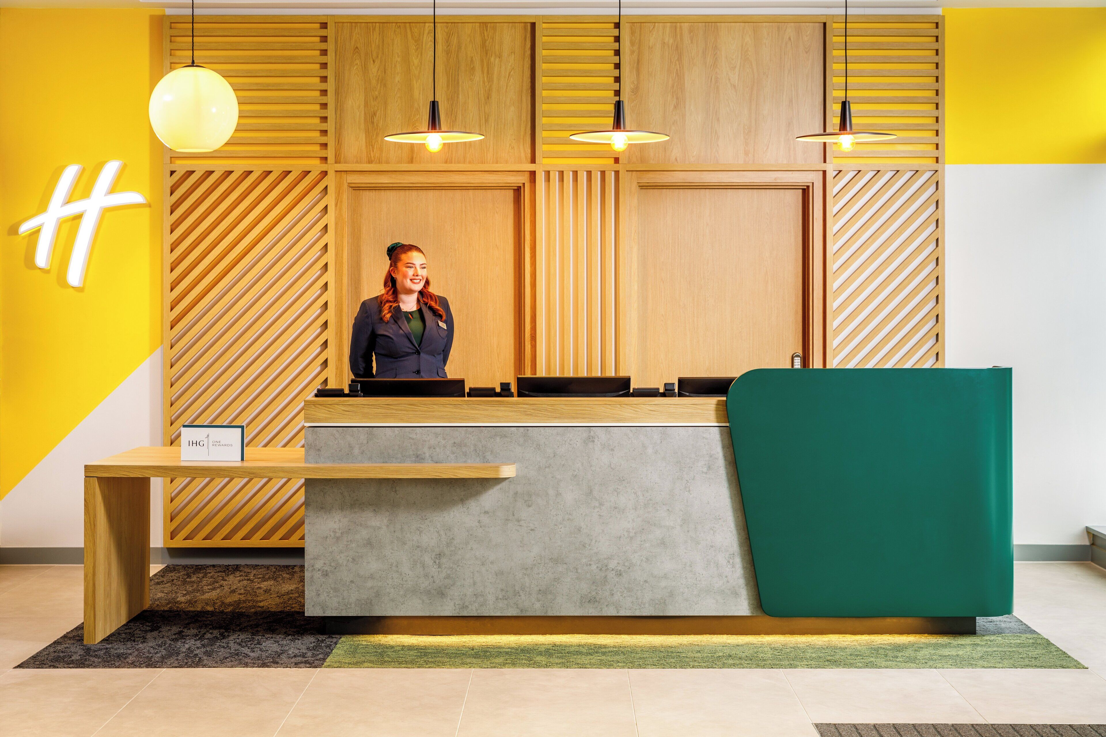 A reception desk with a modern design, featuring a wooden panel wall, a green cushioned seating area, and pendant lighting.