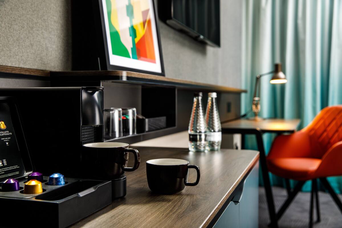 Ein Hotelzimmer mit einem Holztisch, einer schwarzen Kaffeemaschine, einer schwarzen Tasse, einer Wasserflasche und einem orangefarbenen Sessel.