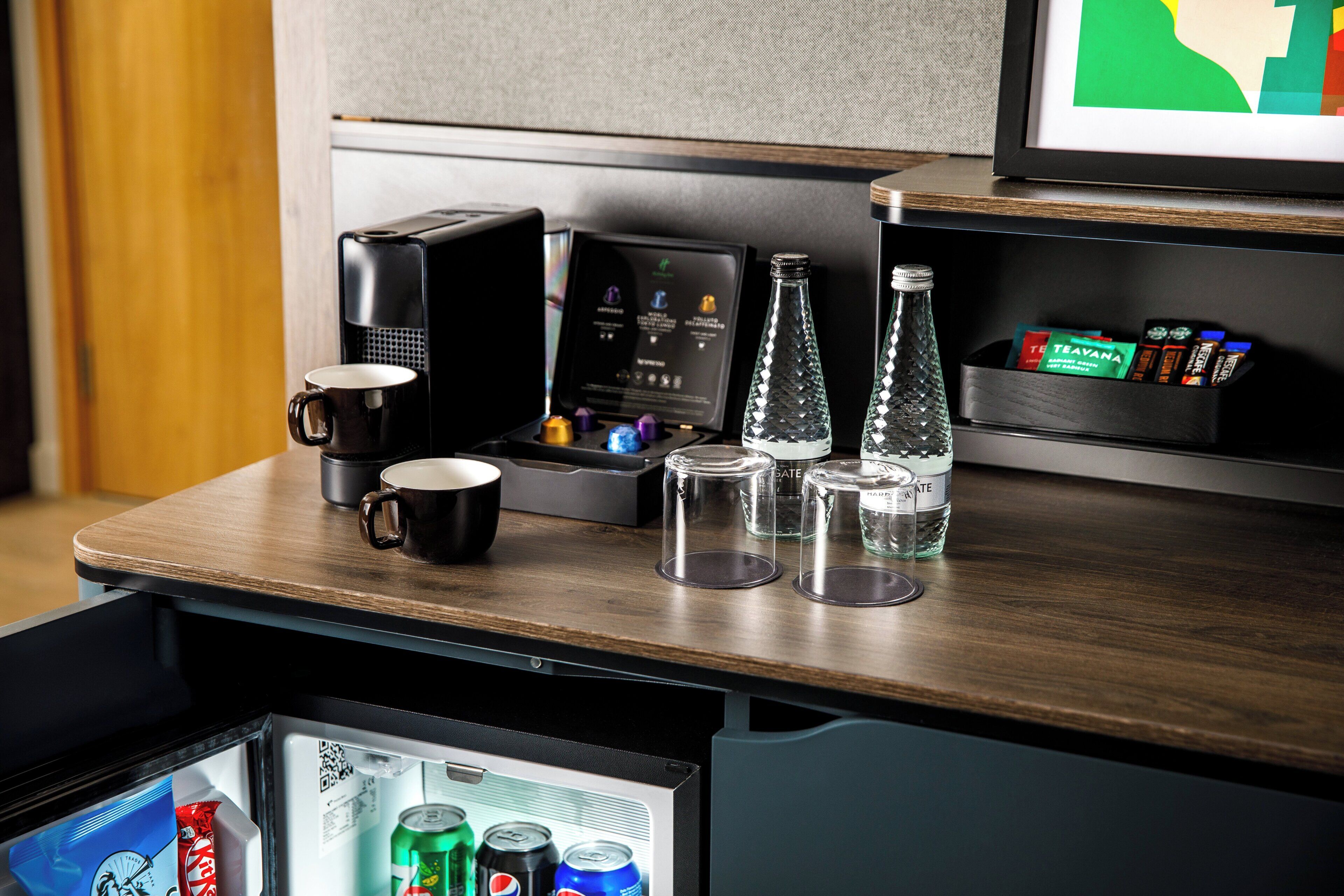 A kitchenette with a coffee maker, a mug, a bottle, and a refrigerator containing beverages.