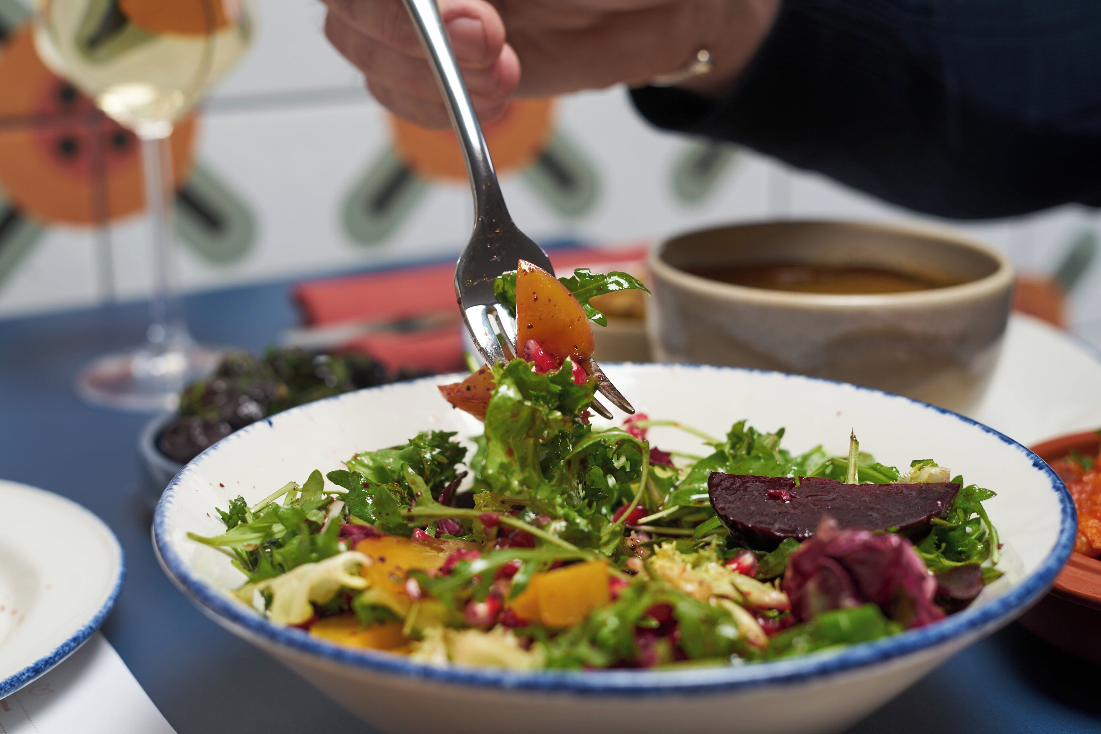Un tazón de ensalada con diferentes hojas verdes y verduras, un tenedor y una copa de vino blanco sobre una mesa.