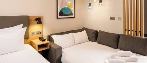 A hotel room with a bed, a sofa, a nightstand, a lamp, and a wall-mounted power outlet.