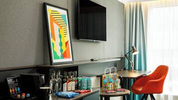 Habitación de hotel con televisor de pantalla plana, un cuadro abstracto enmarcado, un escritorio con cafetera, botellas, latas y un bolso de colores.