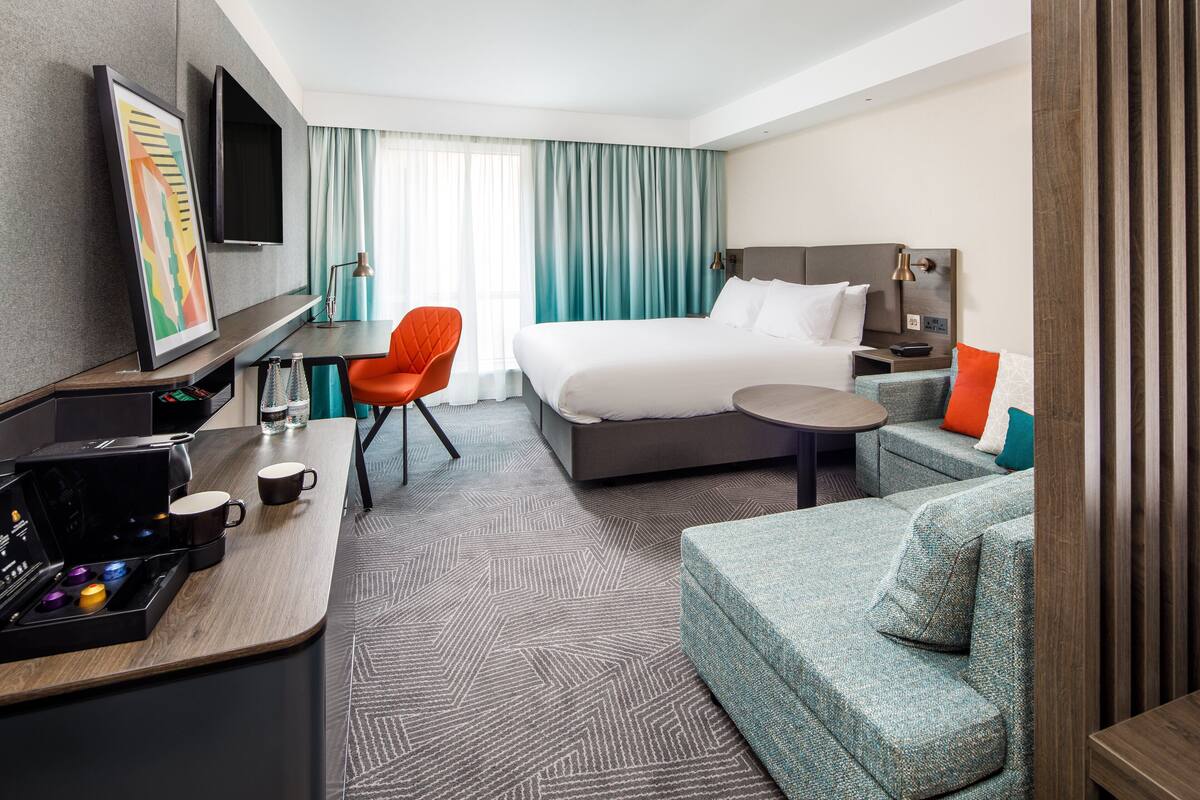 Ein Hotelzimmer mit Bett, Schreibtisch mit Kaffeemaschine, Fernseher und einer Sitzecke mit Sofa und Sessel.