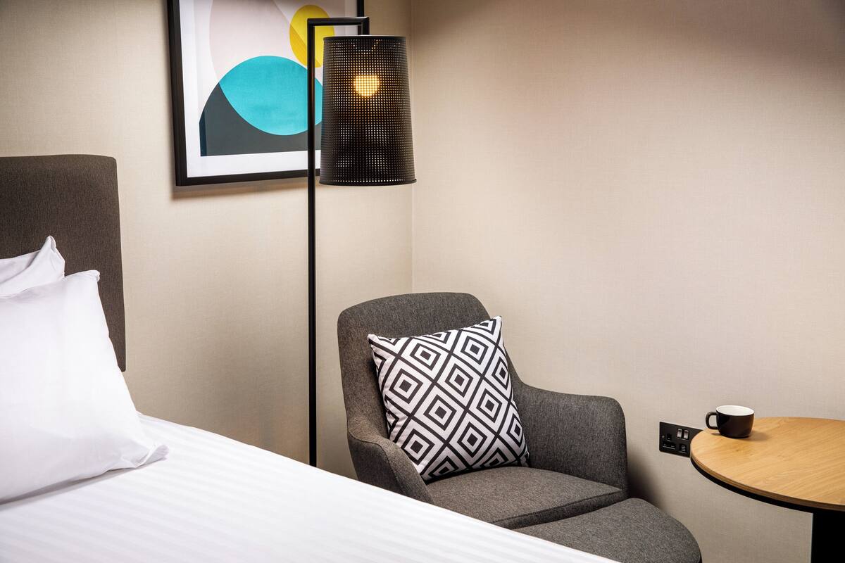 Ein Hotelzimmer mit einem Bett, einem grauen Sessel mit gemustertem Kissen, einem kleinen runden Tisch mit einer Tasse und einer Stehlampe.