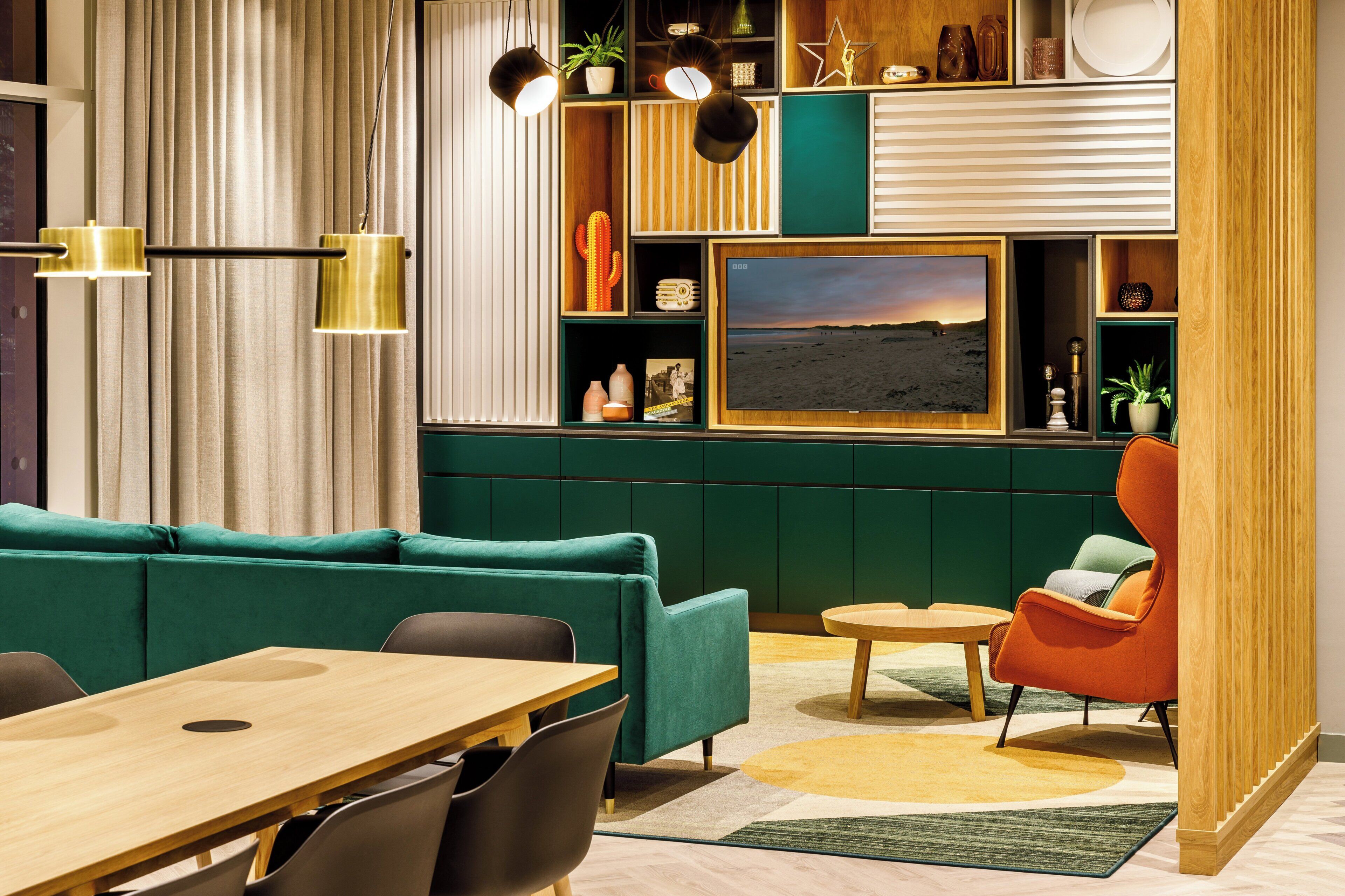 Un salon moderne avec un canapé vert, une table basse en bois et un fauteuil rouge. Un téléviseur est fixé au mur, et une bibliothèque contient divers objets.