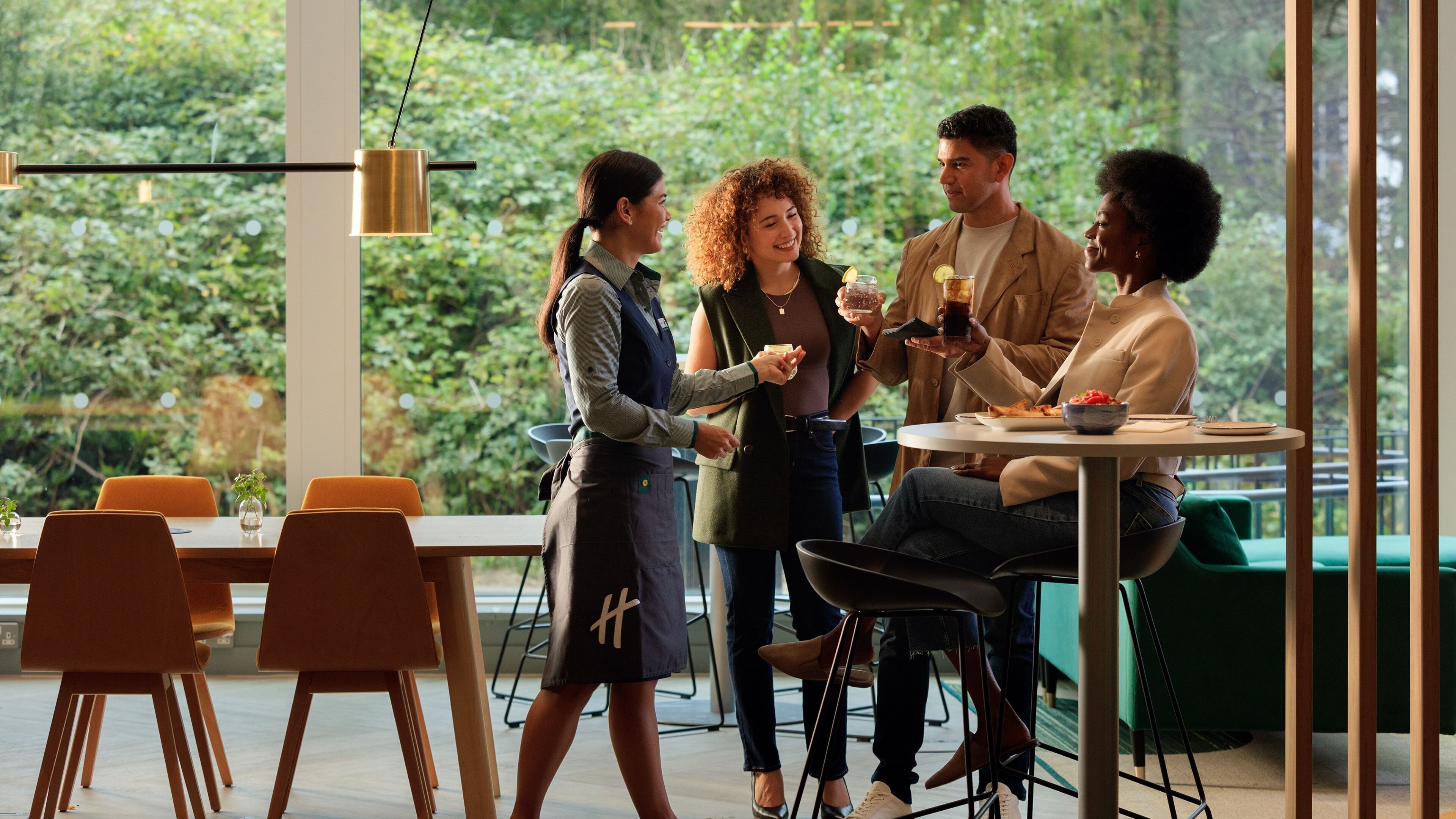 Un groupe de personnes se trouve dans un espace intérieur moderne et bien éclairé, doté de grandes fenêtres, avec une table ronde où sont disposés des plats et des sièges disposés de manière informelle.