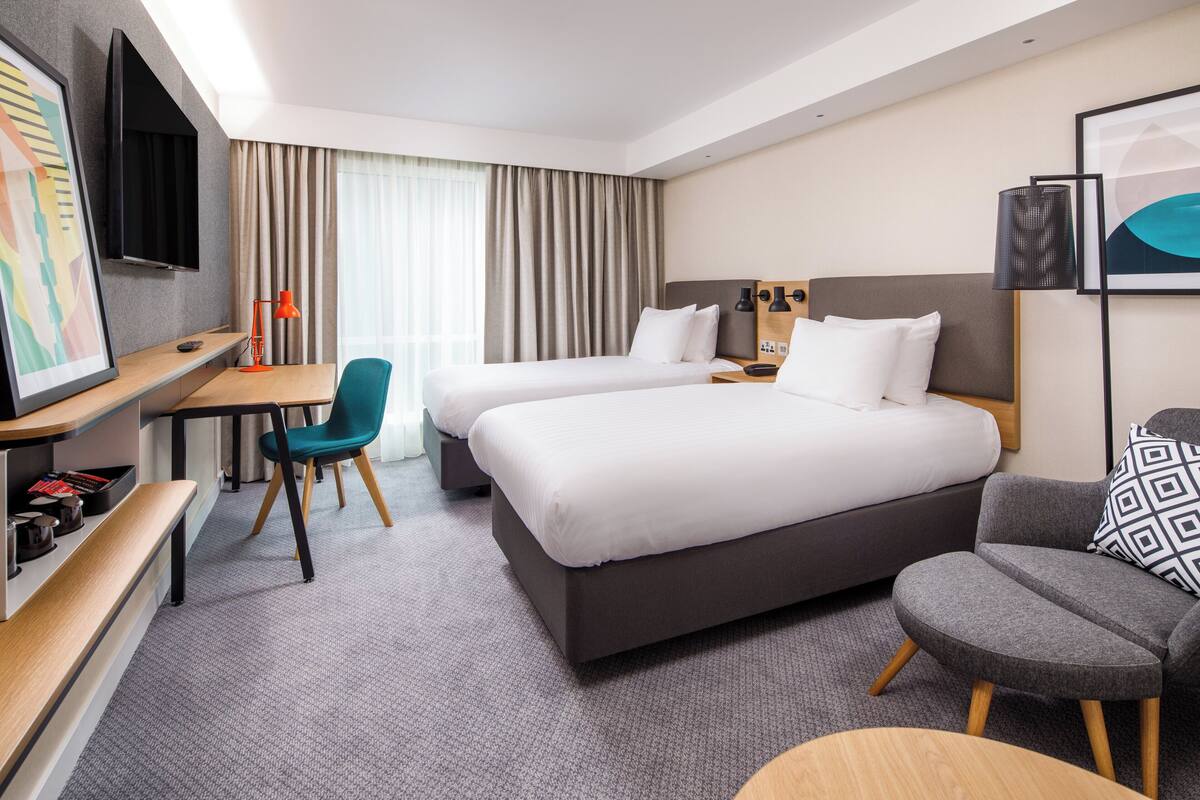 Ein Hotelzimmer mit zwei Betten, einem Schreibtisch mit Stuhl, einem an der Wand montierten Fernseher und einem großen Fenster mit Vorhängen.