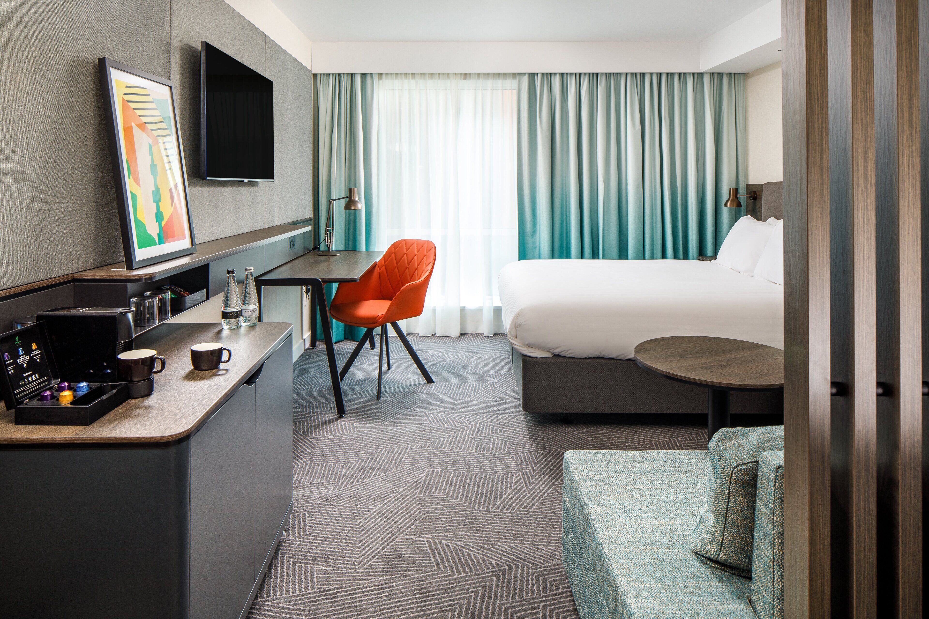 A hotel room with a bed, a desk with a chair, a coffee machine, and a TV on the wall.