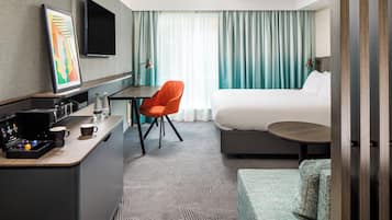 A hotel room with a bed, a desk with a chair, a coffee machine, and a TV on the wall.